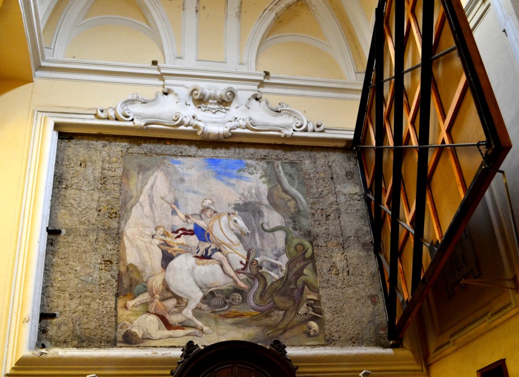 Afresco de Aniello Falcone: São Jorge, em cavalo branco e armado com lança, investe contra um dragão de expressão feroz. Ao fundo, uma princesa foge com véus esvoaçantes, cercada por cadáveres e ossadas humanas. A cena se passa em uma paisagem rochosa, com céu em movimento e vegetação retorcida, revelando um equilíbrio entre teatralidade e contenção trágica.