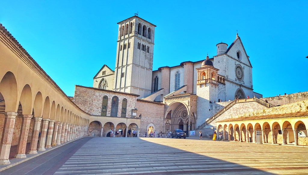 Vista ampla da esplanada inferior da Basílica de São Francisco em Assis. À esquerda e à direita, galerias com arcos de pedra criam uma moldura simétrica para o espaço central. Ao fundo, a fachada e a torre da basílica se erguem em tons claros sob o céu azul. A cena está banhada por luz do fim da tarde, com alguns visitantes dispersos.